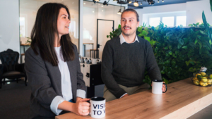 Zwei Experten sitzen an einem Holztisch in einem modernen Büro und unterhalten sich entspannt bei Kaffee.