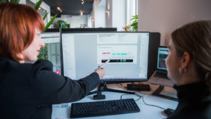 Zwei Frauen arbeiten gemeinsam an einem Schreibtisch in einem modernen Büro; eine zeigt mit einem Stift auf Inhalte auf einem großen Monitor, während die andere aufmerksam zusieht, daneben steht ein aufgeklappter Laptop.