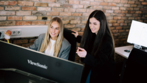 Zwei Frauen sitzen gemeinsam an einem Schreibtisch vor einem großen Monitor und lächeln, während sie sich etwas auf dem Bildschirm ansehen; eine zeigt mit der Hand darauf, im Hintergrund ist eine Backsteinwand zu sehen.